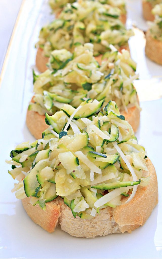 Slices of toasted bread become zucchini crostini, topped with a savory mixture of shredded zucchini and cheese, arranged neatly on a white rectangular platter.