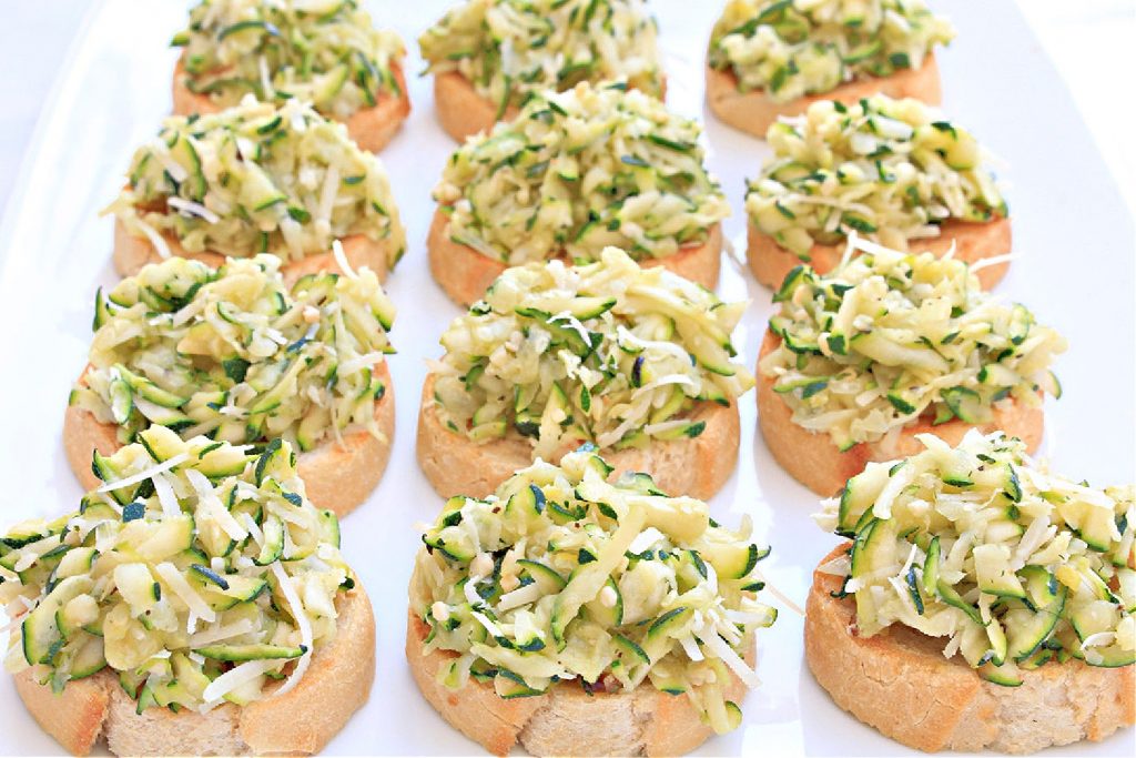 Slices of baguette topped with a zucchini crostini mixture of shredded zucchini and cheese are arranged in rows on a white platter.