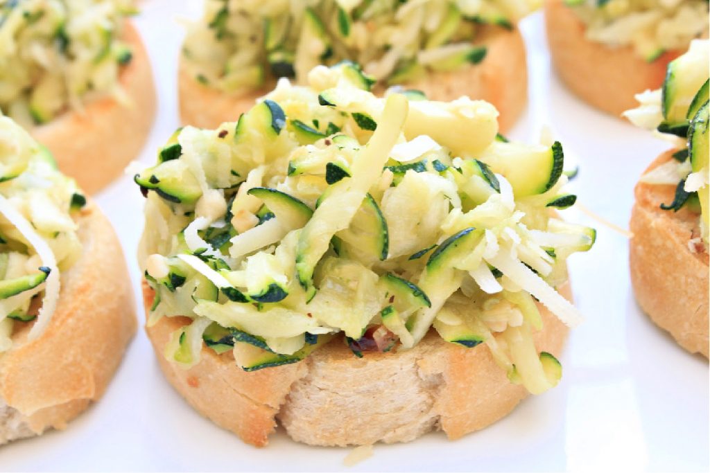 Slices of baguette topped with a generous serving of shredded zucchini and cheese create a delicious zucchini crostini, arranged on a white plate. The zucchini appears fresh and lightly seasoned.