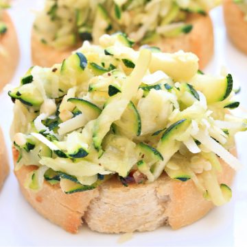 Slices of bread topped with a generous portion of shredded zucchini and cheese create savory zucchini crostini, arranged on a white surface. The zucchini appears fresh and lightly seasoned.