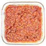 A glass baking dish filled with vegan BBQ baked beans in a rich, thick reddish-brown sauce, viewed from above on a white background.