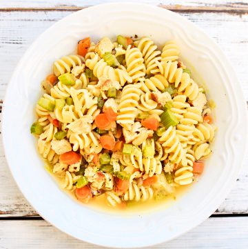 A white bowl filled with rotini pasta, vegan chicken, carrots, and celery in a light broth, set on a rustic white wooden surface.