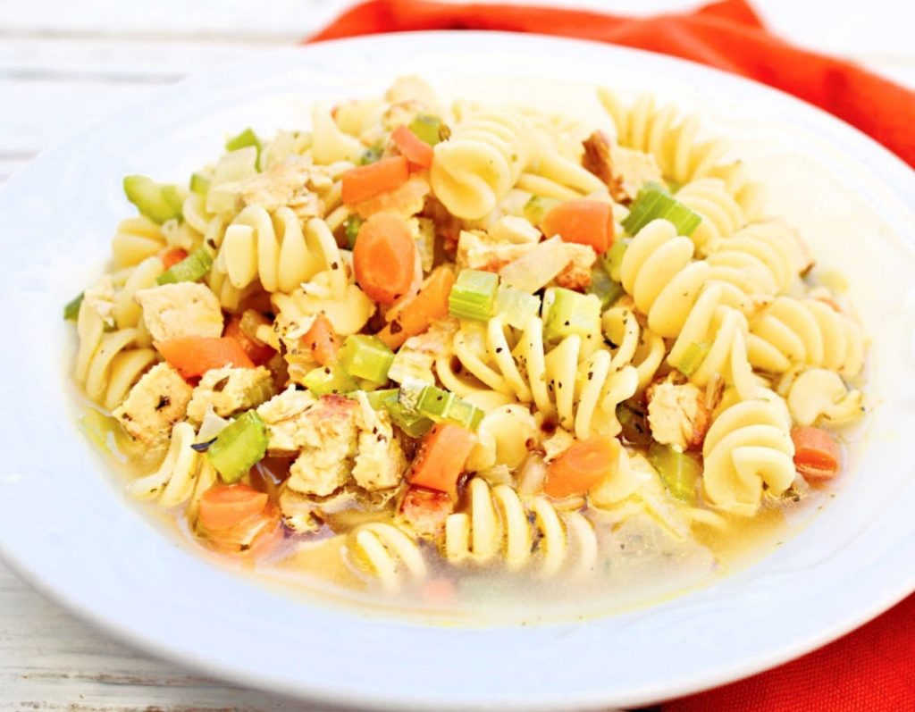 A white plate filled with rotini pasta soup, featuring chunks of vegan chicken, sliced carrots, celery, and a light broth, set on a white table with an orange napkin in the background.