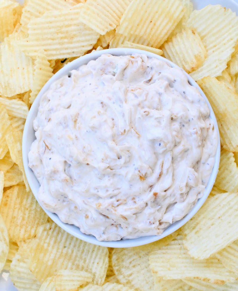 A bowl of creamy French onion dip sits in the center of a plate surrounded by ridged potato chips.