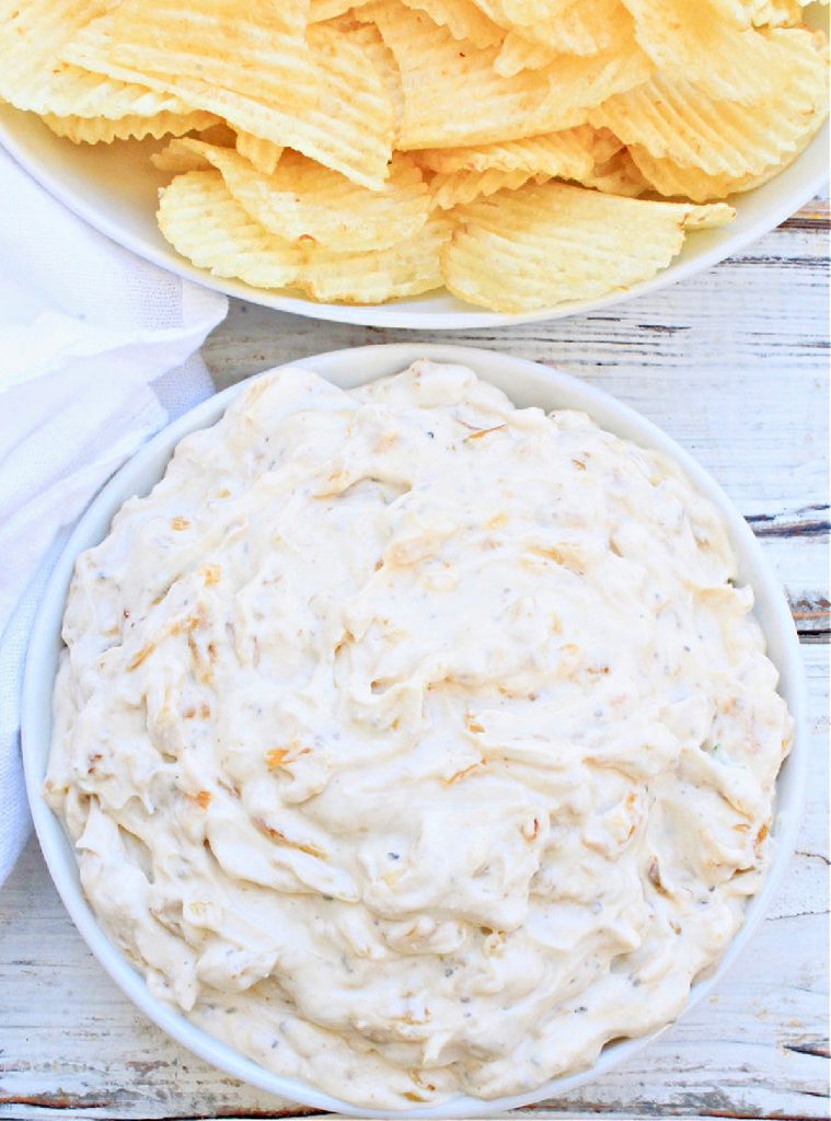 A bowl of creamy french onion dip sits next to a bowl of ridged potato chips on a white wooden surface. A white napkin is partially visible beside the bowls, making this French Onion Dip Recipe perfect for snacking.
