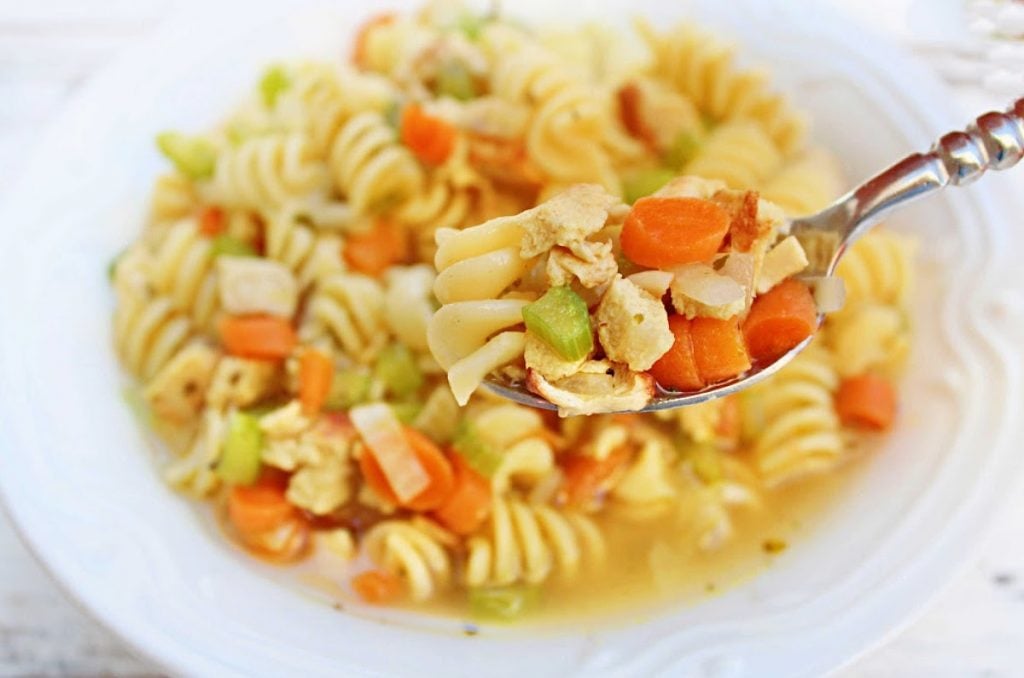 A close-up of a bowl of vegan chicken noodle soup with rotini pasta, diced carrots, celery, and plant-based chicken. A spoon lifts a portion from the bowl, showcasing the colorful ingredients.