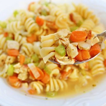 A close-up of a bowl of vegan chicken noodle soup with rotini pasta, diced carrots, celery, and plant-based chicken. A spoon lifts a portion from the bowl, showcasing the colorful ingredients.