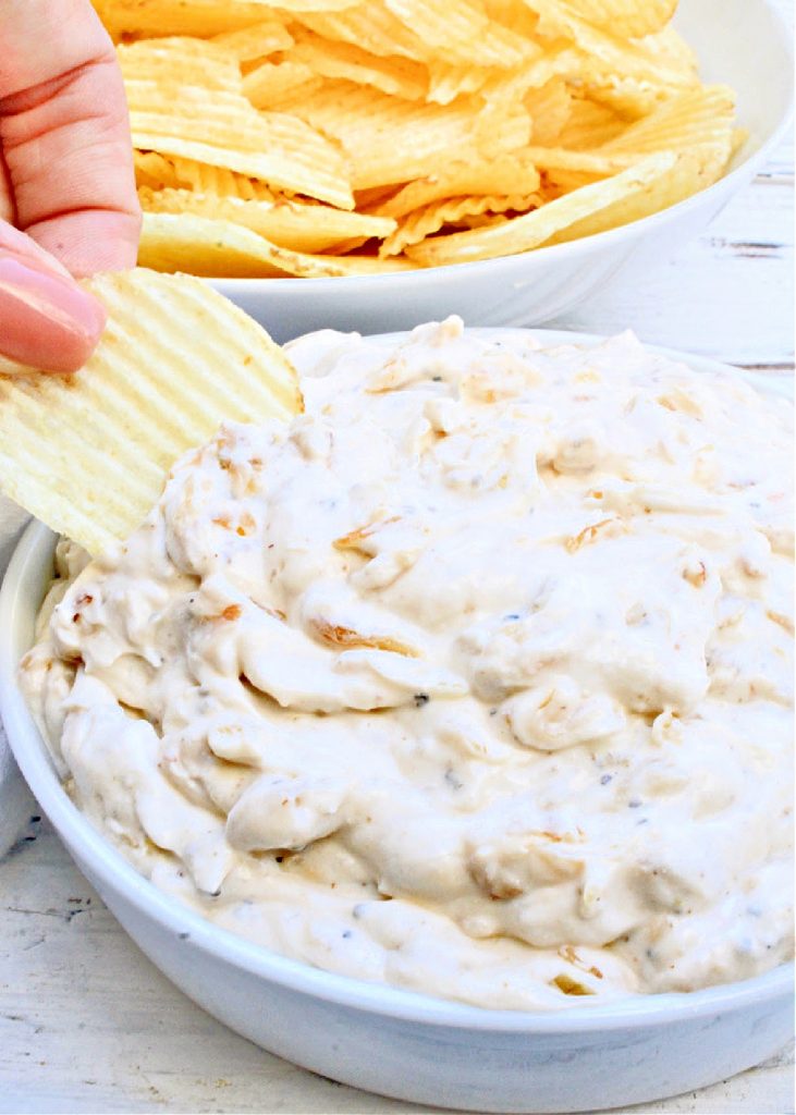 A hand dips a ridged potato chip into a bowl of creamy french onion dip, with another bowl of potato chips in the background on a white wooden surface.