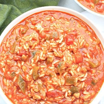 A white bowl filled with hearty stuffed pepper soup—made with rice, ground meat, tomatoes, bell peppers, and broth; a green cloth is nearby and another bowl of stuffed pepper soup is partially visible in the background.