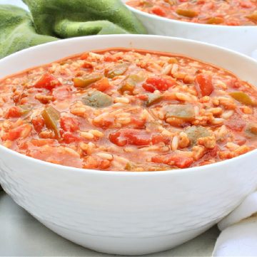 A white bowl filled with hearty stuffed pepper soup—a tomato-based broth with rice, chopped tomatoes, green peppers, and other vegetables—sits beside green and white cloth napkins.