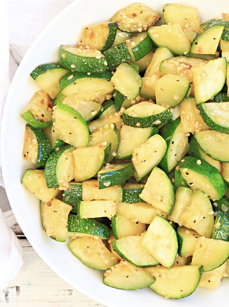 A close-up of a white bowl filled with garlic butter zucchini slices, sautéed to perfection and topped with sesame seeds and spices.