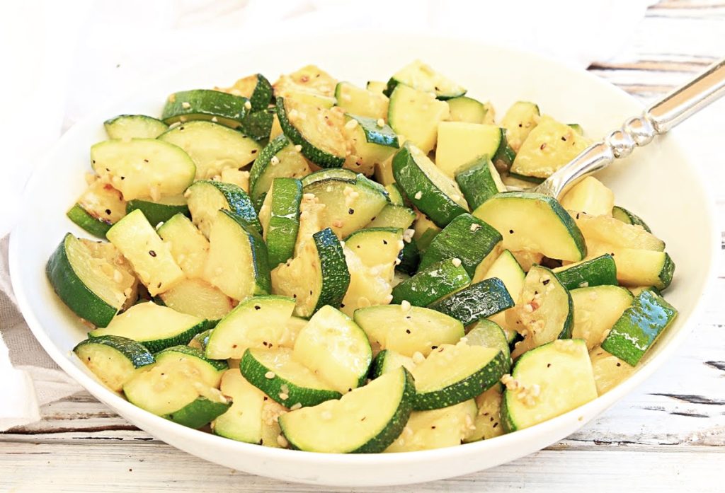 A white bowl filled with garlic butter zucchini sautéed slices, lightly seasoned and garnished with sesame seeds, with a silver spoon resting inside. The background is a rustic white wood surface.