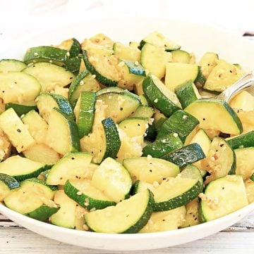 A white bowl filled with garlic butter zucchini slices, lightly seasoned and sautéed, with a silver serving spoon on the side. The background features a rustic white wooden surface.
