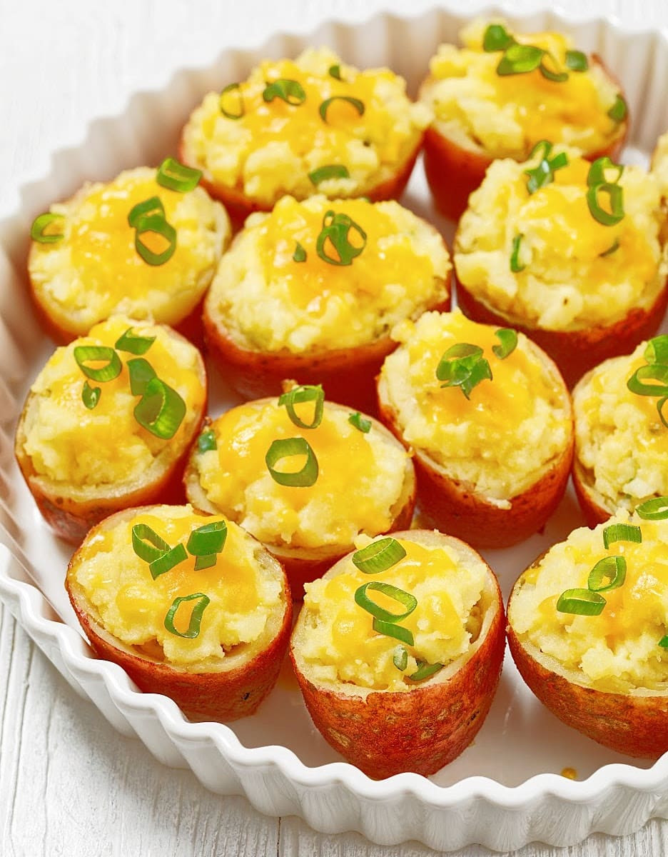 A white dish filled with stuffed baby red potatoes, each half topped with melted cheddar cheese and chopped green onions, arranged neatly in rows.