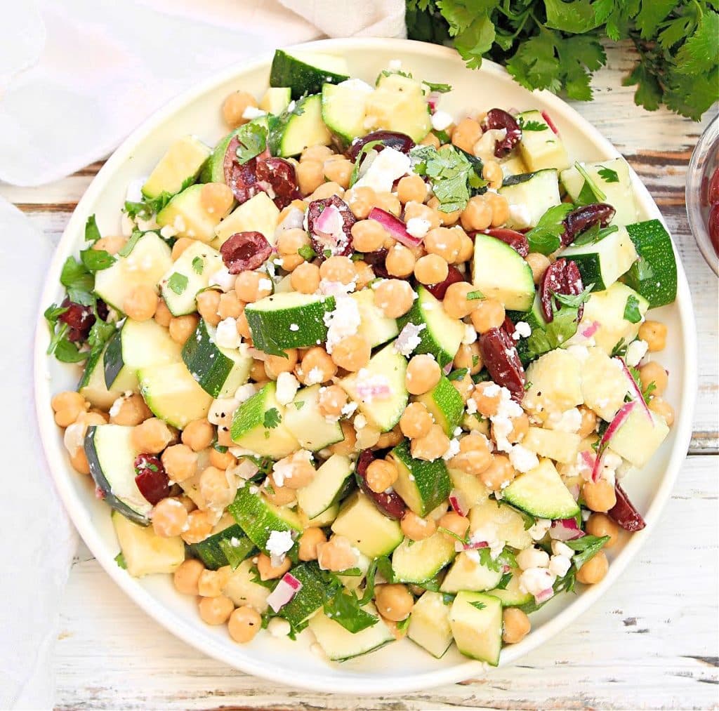 A plate of chickpea salad with chopped cucumber, kalamata olives, red onion, feta cheese, and fresh herbs, reminiscent of a Greek zucchini salad, arranged on a white plate atop a rustic white wooden surface.
