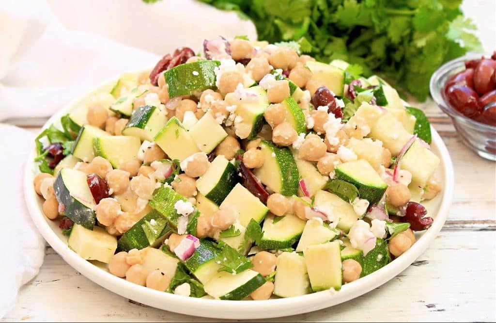 A plate of fresh summer zucchini salad with chickpeas, red onion, feta cheese, parsley, and kidney beans sits on a white wooden table with a bowl of grapes and cilantro in the background.