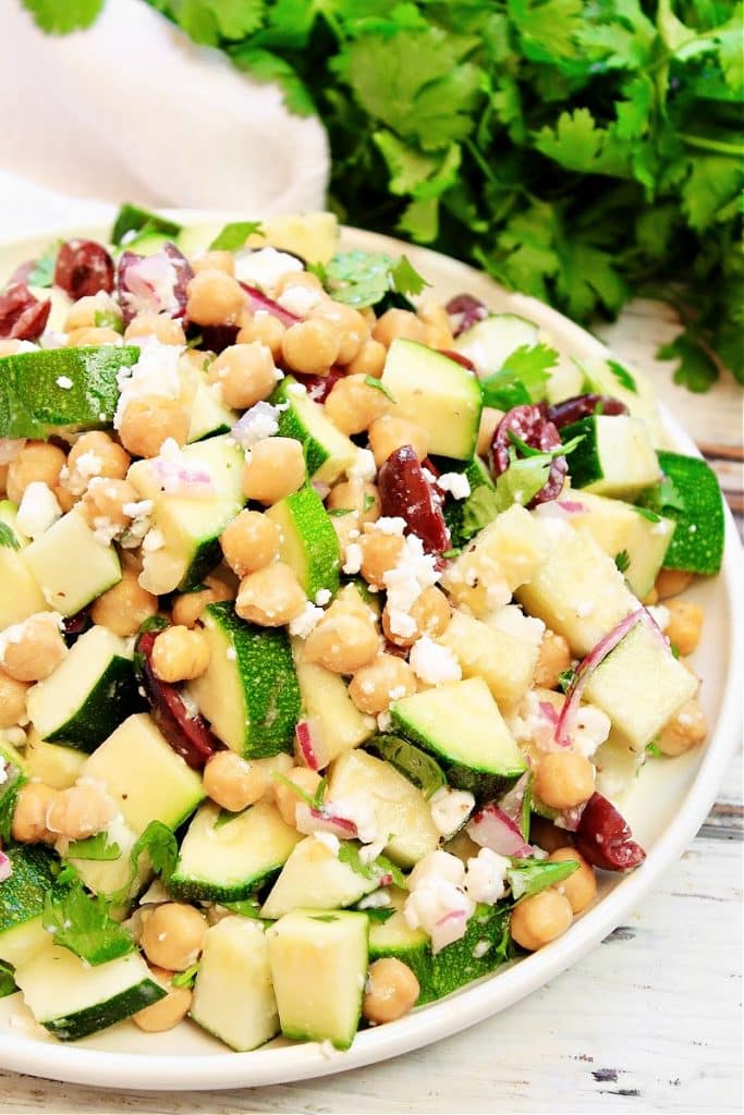 A vibrant Greek zucchini salad with chopped zucchini, chickpeas, kalamata olives, red onions, feta cheese, and fresh cilantro, served on a white plate with extra cilantro in the background.