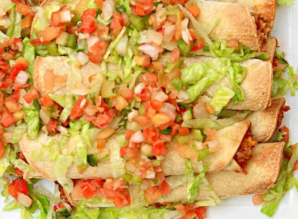 A plate of bean and cheese taquitos, baked and rolled, topped with shredded lettuce and chopped tomato salsa, arranged closely together on a white platter.