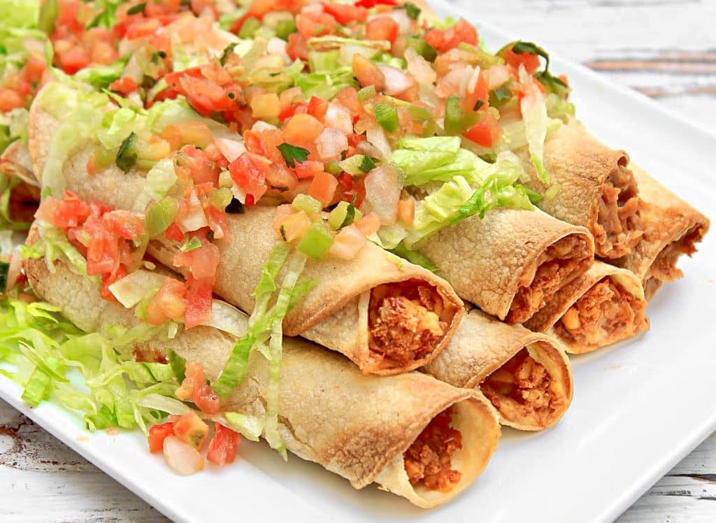 A plate of crispy chicken taquitos and bean and cheese taquitos topped with chopped lettuce, diced tomatoes, onions, and green peppers, served on a white rectangular dish.