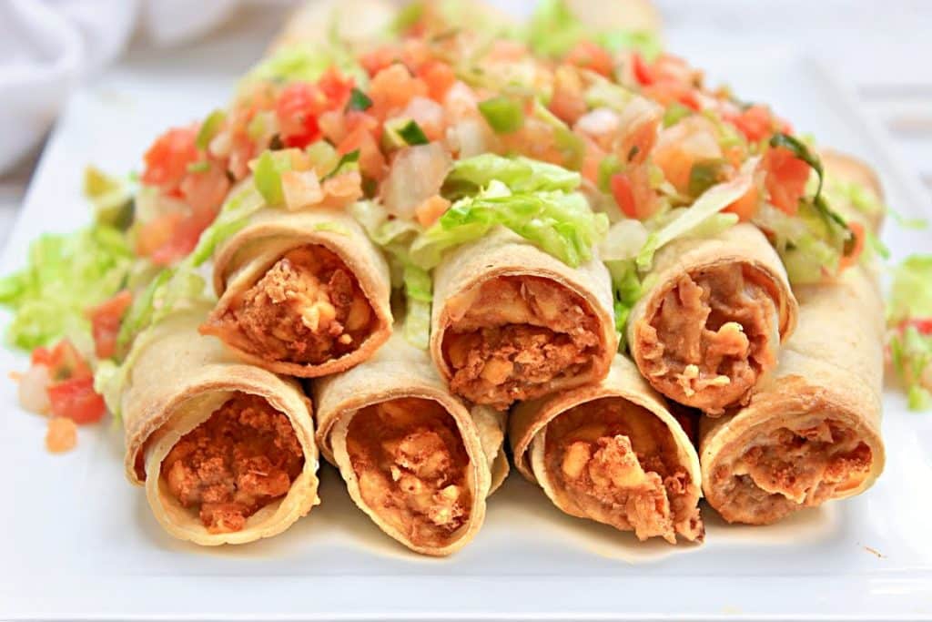 Six crispy rolled tacos, including our savory bean and cheese taquitos filled with seasoned meat, are arranged on a white plate, topped with shredded lettuce and fresh pico de gallo salsa.