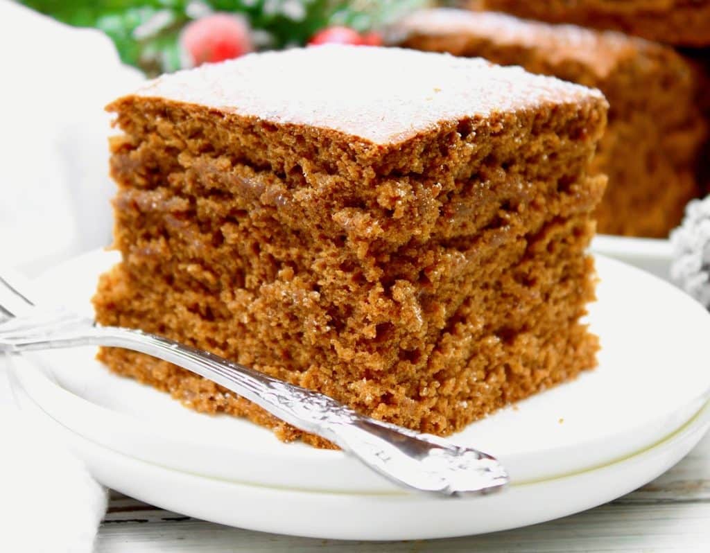 A close-up of a thick slice of moist vegan gingerbread cake on a white plate with a silver fork, topped with a light dusting of powdered sugar. The background is softly blurred with hints of greenery and red berries.