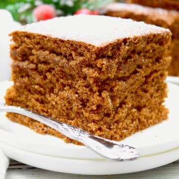 A close-up of a thick, square slice of vegan gingerbread cake on a white plate with a silver fork, showing its moist, fluffy texture. The background is softly blurred with festive greenery and red berries.