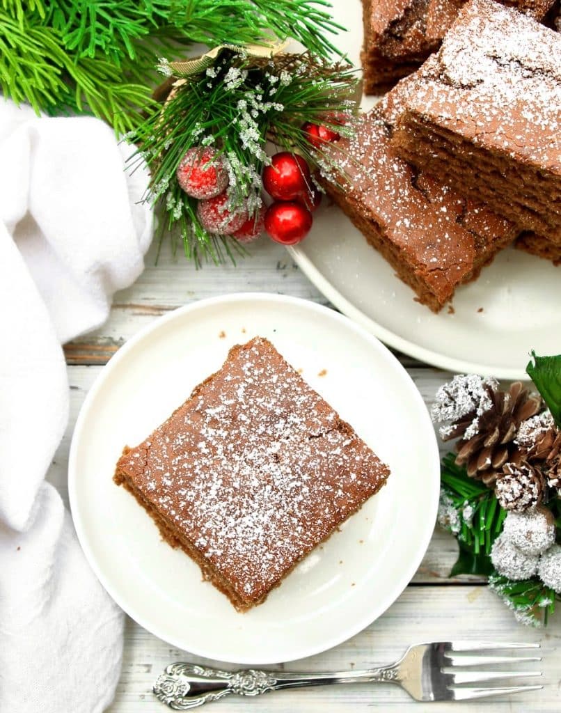 A square piece of vegan gingerbread cake dusted with powdered sugar sits on a white plate, surrounded by festive pine branches, red berries, pine cones, and more cake slices on a platter. A silver fork rests nearby.