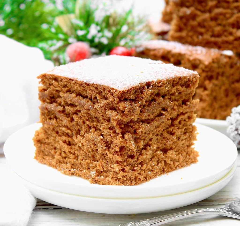 A close-up of a square slice of vegan gingerbread cake on a white plate. The cake has a fluffy, moist texture and is lightly dusted with powdered sugar. There are festive decorations and greenery blurred in the background.