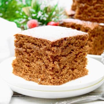 A close-up of a square slice of vegan gingerbread cake on a white plate. The cake has a fluffy, moist texture and is lightly dusted with powdered sugar. There are festive decorations and greenery blurred in the background.