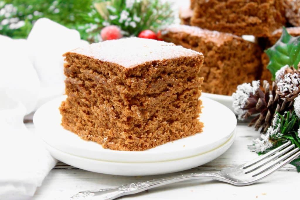 A slice of moist vegan gingerbread cake sits on a white plate, with a fork nearby. Festive greenery, pinecones, and red berries in the background evoke a cozy holiday atmosphere.