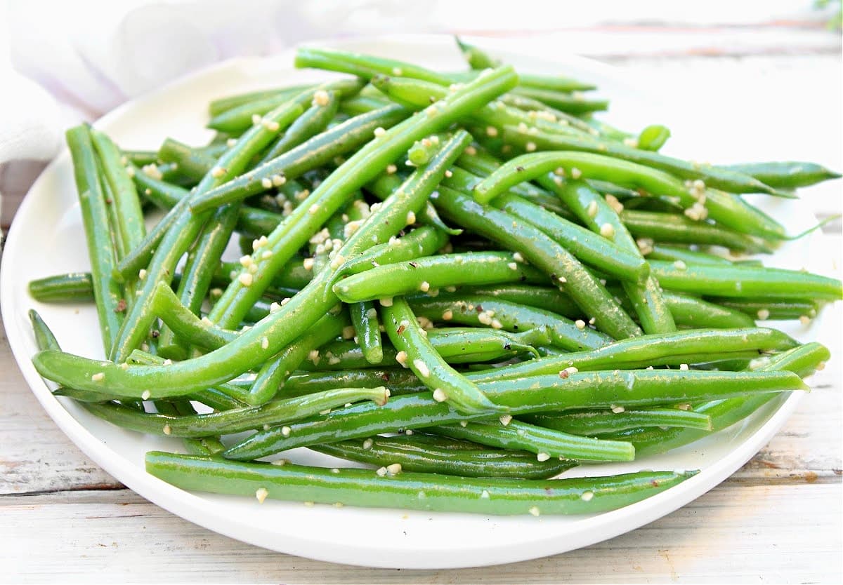 A white plate piled with bright green garlic green beans, garnished with minced garlic and seasonings, sits on a light wooden surface.