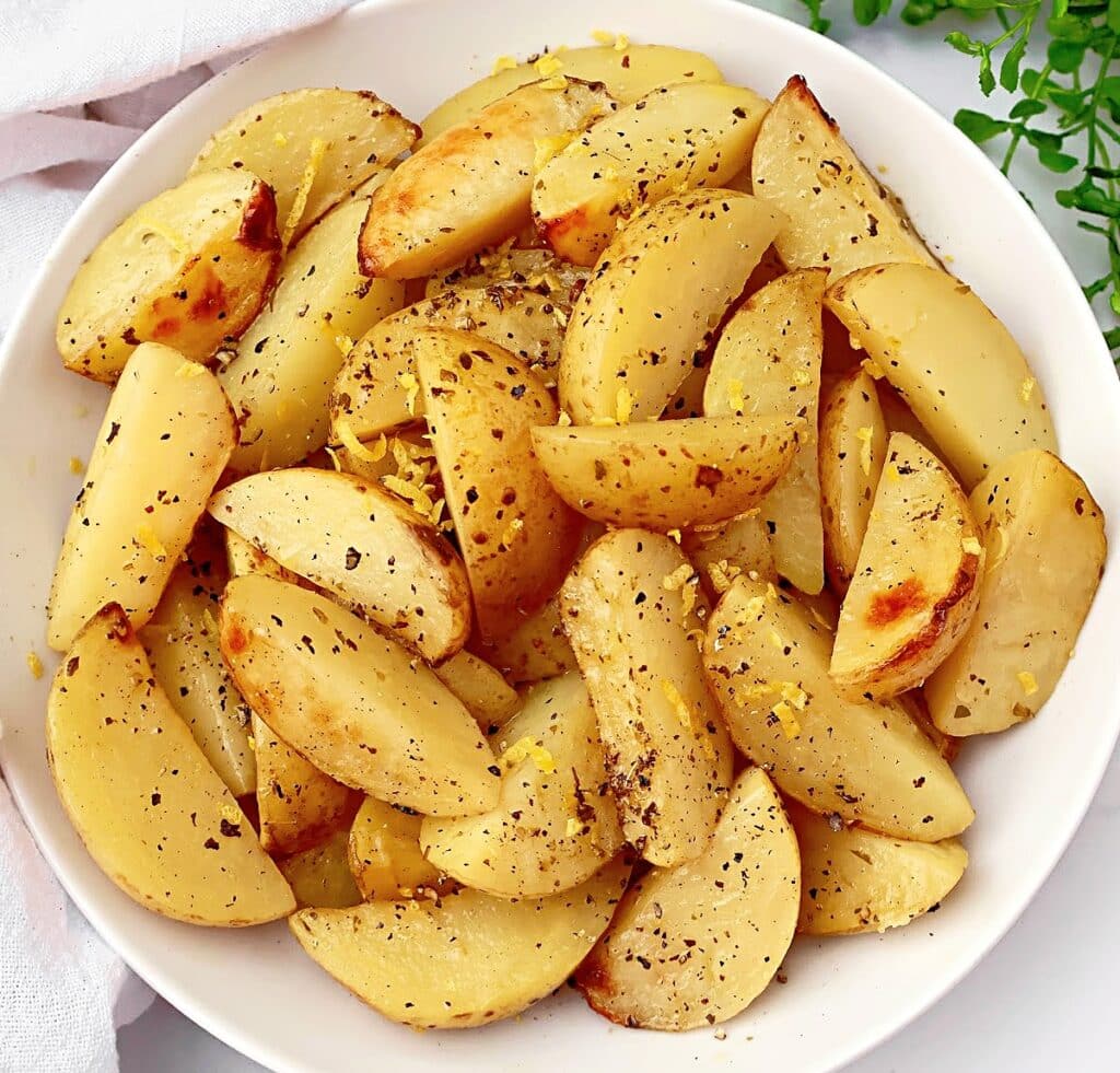 A white bowl filled with golden-brown lemon roasted potatoes, seasoned with herbs and black pepper, and garnished with fresh lemon zest.