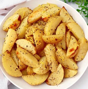 A white bowl filled with lemon roasted potatoes, seasoned with herbs and black pepper, garnished with lemon zest. The bowl is set on a white surface with a green plant visible in the background.
