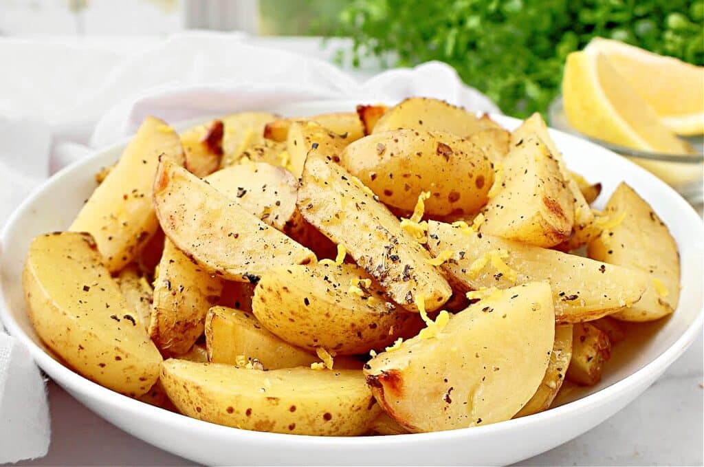A white bowl filled with lemon roasted potatoes, seasoned with black pepper and lemon zest, sits on a table. In the background, there are lemon slices and green herbs.