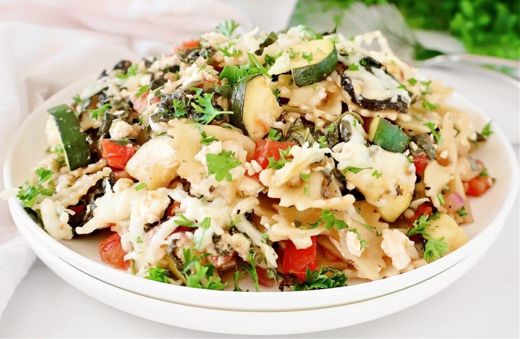A plate of garden vegetable pasta with bowtie pasta, zucchini, tomatoes, herbs, leafy greens, melted cheese, and fresh parsley. This vibrant summer pasta recipe is beautifully garnished and served on a white plate.