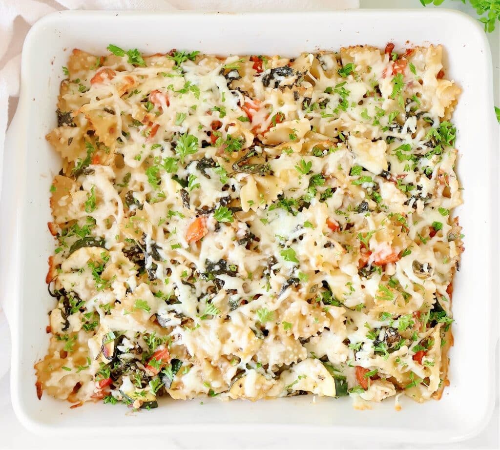 A baked garden vegetable pasta casserole in a white dish, topped with melted cheese, chopped herbs, tomatoes, and vegetables. This summer pasta recipe has a golden, bubbly appearance and is garnished with fresh green parsley.