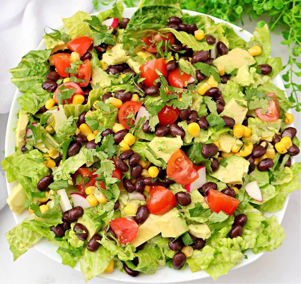A fresh Mexican chopped salad featuring romaine lettuce, black beans, corn, diced avocado, cherry tomatoes, red onion, and cilantro, all served in a white bowl.