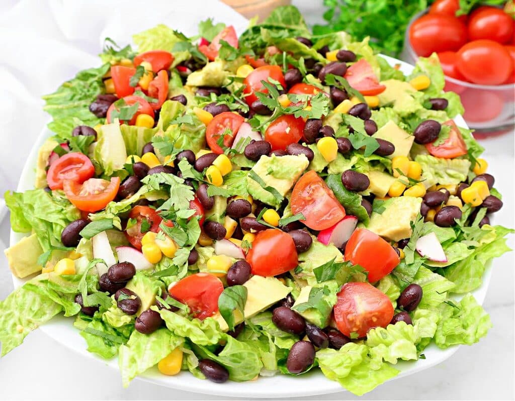 A vibrant Mexican chopped salad with romaine lettuce, black beans, corn, avocado, radishes, cherry tomatoes, and cilantro is served in a white bowl. A bowl of tomatoes appears in the background.