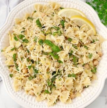 A white bowl filled with farfalle pasta salad, garnished with chopped herbs, shredded cheese, and two lemon wedges. The salad includes chickpeas, herbs, and is seasoned with black pepper. A bunch of fresh herbs is in the background.