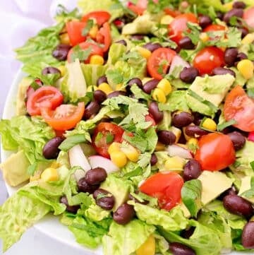 A fresh Mexican chopped salad with romaine lettuce, cherry tomatoes, black beans, corn, avocado chunks, diced onions, and cilantro served in a white bowl.