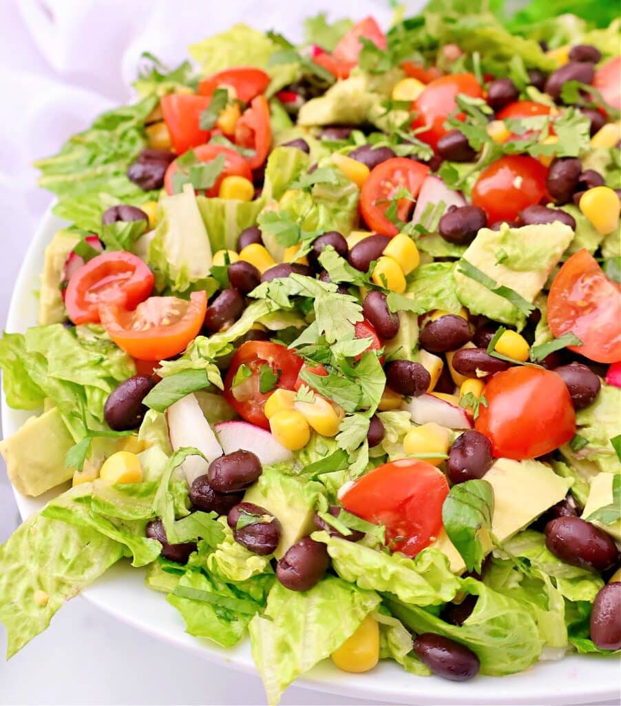 A fresh Mexican chopped salad with romaine lettuce, cherry tomatoes, black beans, corn, avocado chunks, diced onions, and cilantro served in a white bowl.