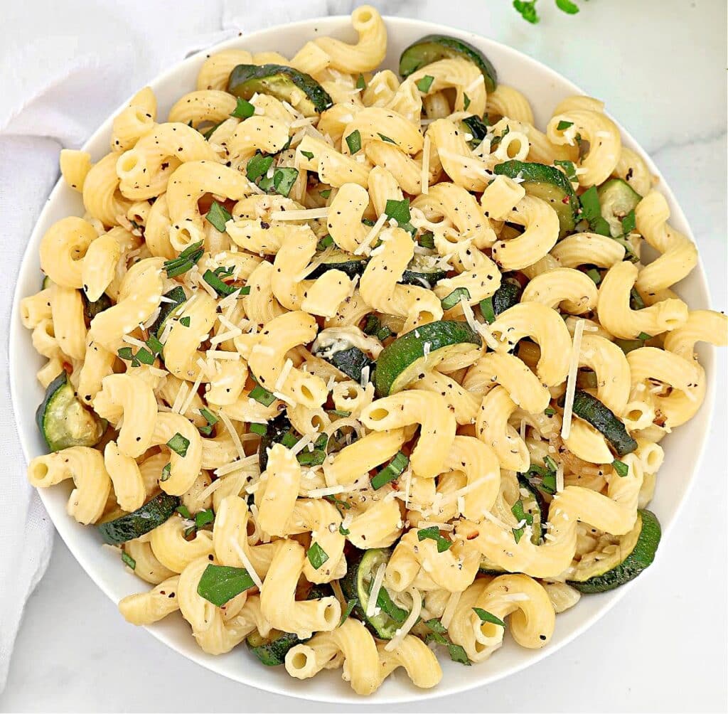 A white bowl filled with pasta spirals, sliced zucchini, grated cheese, chopped herbs, and cracked black pepper, placed on a white surface.