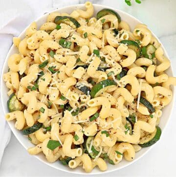 A white bowl filled with pasta spirals, sliced zucchini, grated cheese, chopped herbs, and cracked black pepper, placed on a white surface.