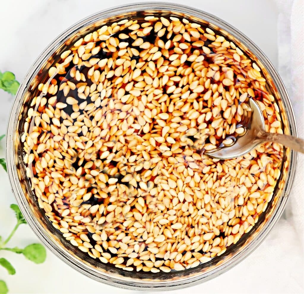 A glass bowl filled with a dark liquid and floating sesame seeds, with a metal spoon partially submerged on the right side. A green plant and a light cloth are visible in the background.