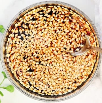 A glass bowl filled with a dark liquid and floating sesame seeds, with a metal spoon partially submerged on the right side. A green plant and a light cloth are visible in the background.