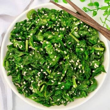 A bowl of cooked spinach garnished with white sesame seeds, with a pair of wooden chopsticks resting on the rim. Fresh spinach leaves and a white napkin are visible in the background.