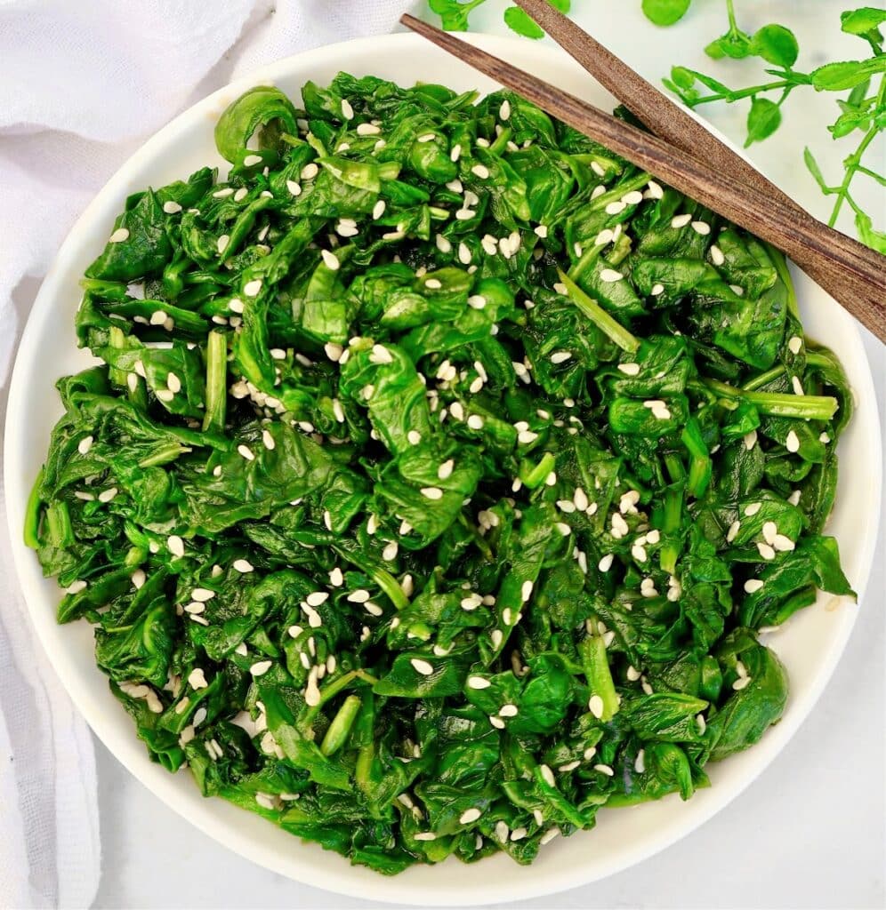 A bowl of cooked spinach garnished with white sesame seeds, with a pair of wooden chopsticks resting on the rim. Fresh spinach leaves and a white napkin are visible in the background.