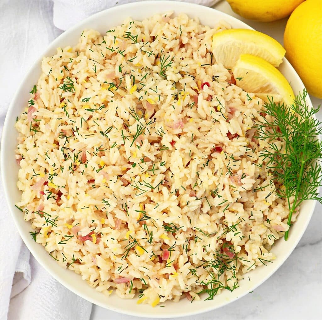 A bowl of lemon rice garnished with fresh dill and lemon slices, viewed from above. The rice is mixed with herbs and small bits of vegetables, creating a colorful and appetizing presentation.