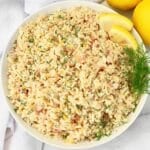 A bowl of lemon rice garnished with fresh dill and lemon slices, viewed from above. The rice is mixed with herbs and small bits of vegetables, creating a colorful and appetizing presentation.