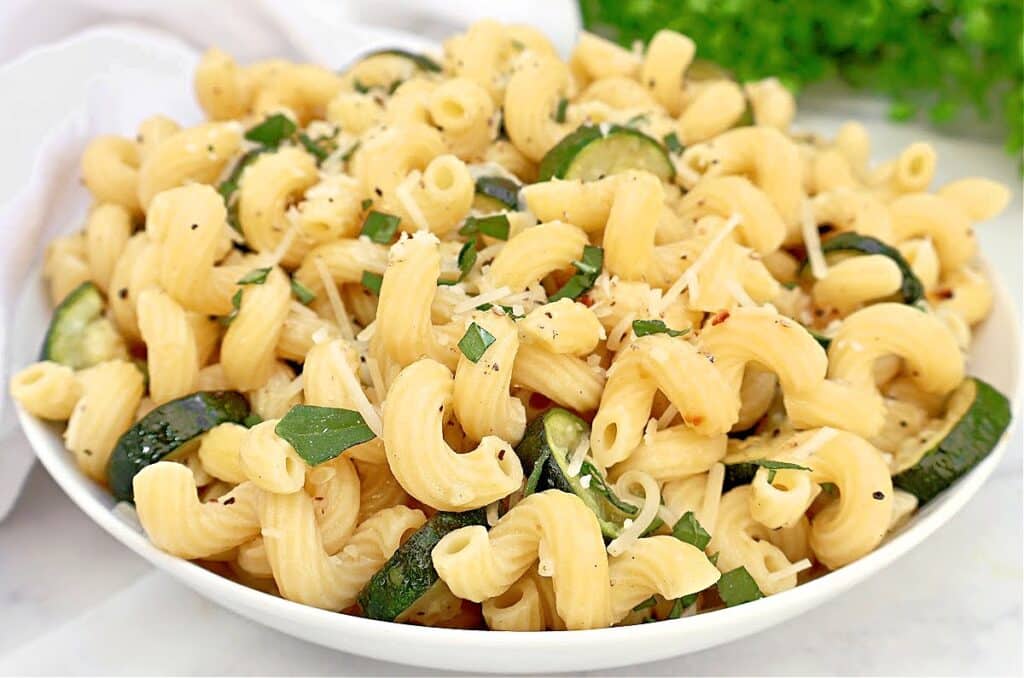 A bowl of cooked cavatappi pasta mixed with slices of zucchini, topped with grated cheese, chopped basil, and cracked black pepper.